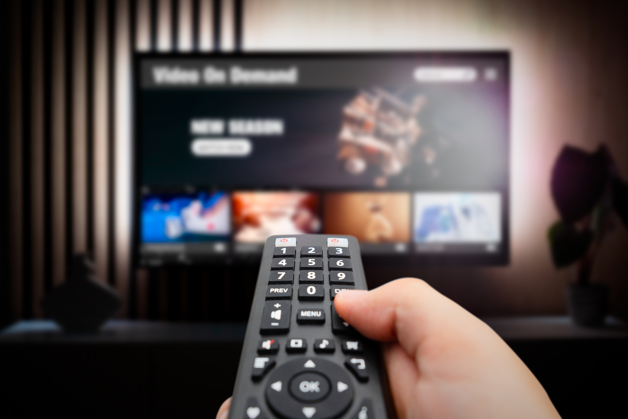 A hand holding a remote control is pointed at a TV screen displaying a video-on-demand menu showing various media options