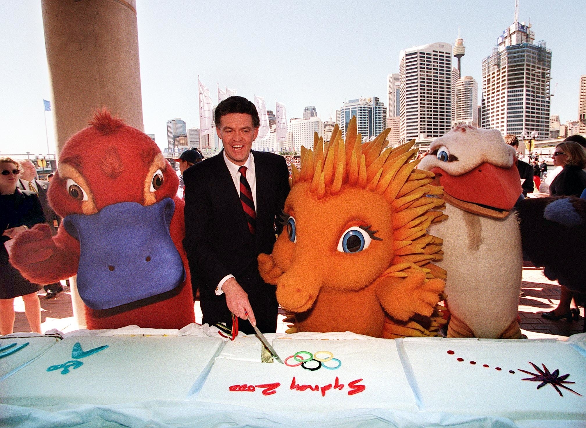 A man in a suit stands with Olympic mascots Syd the platypus, Olly the kookaburra, and Millie the echidna, cutting a large cake with Sydney's skyline in the background