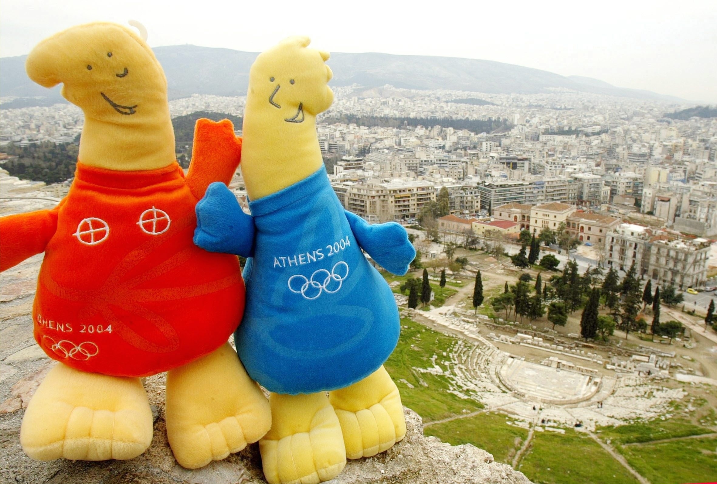 Two plush mascots from the Athens 2004 Olympics, dressed in orange and blue outfits, are posed together with a view of a cityscape in the background