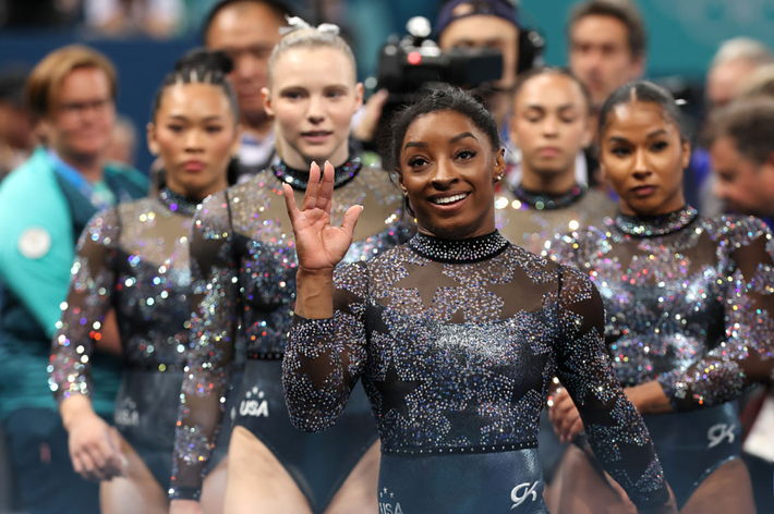 Simone Biles and fellow USA gymnasts in sparkling leotards, smiling and waving at a gymnastics event