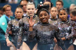 Simone Biles and fellow USA gymnasts in sparkling leotards, smiling and waving at a gymnastics event