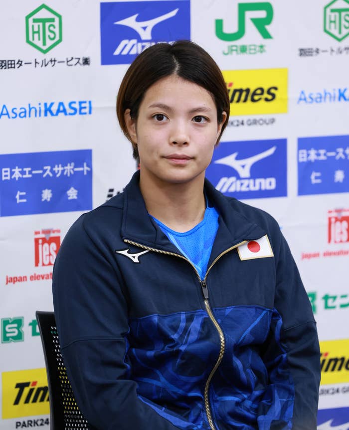 女性がインタビューの背景の前で座っている。青いジャケットを着ており、胸に日本の国旗が付いている。