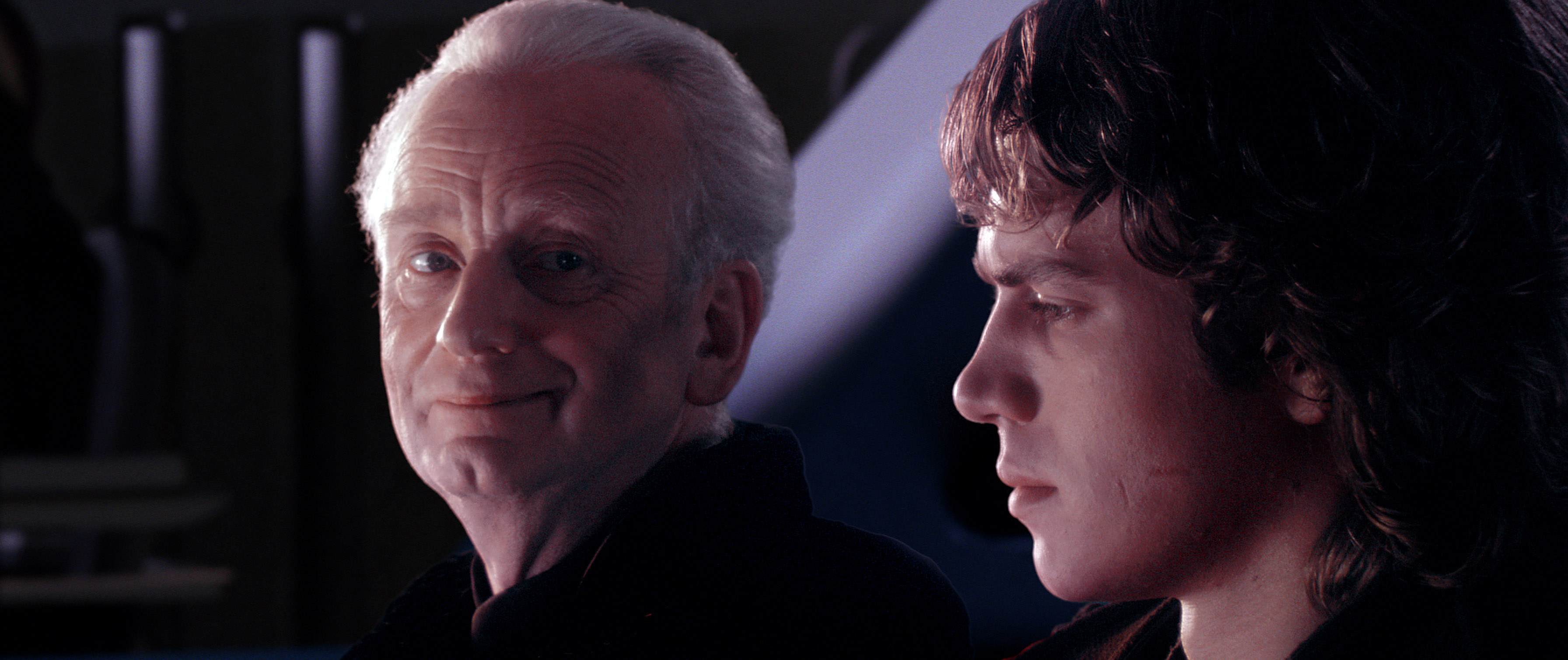 Ian McDiarmid, as Emperor Palpatine, smiles while sitting beside Hayden Christensen, as Anakin Skywalker, from a scene in a Star Wars movie