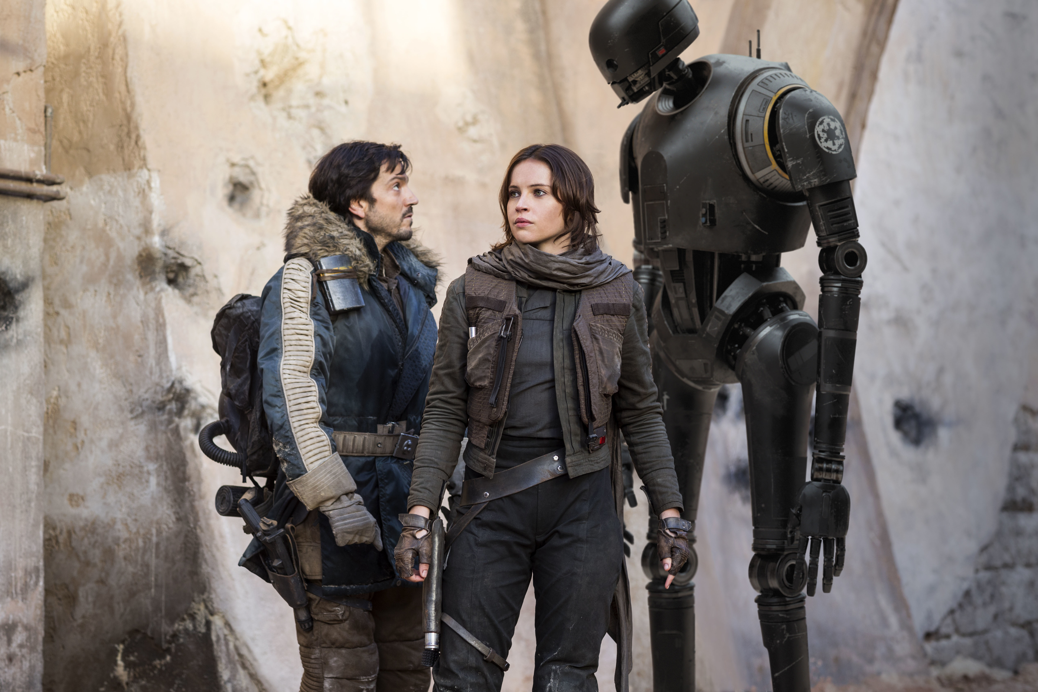 Diego Luna, Felicity Jones, and the droid K-2SO stand together in rugged, tactical outfits in a scene from a movie