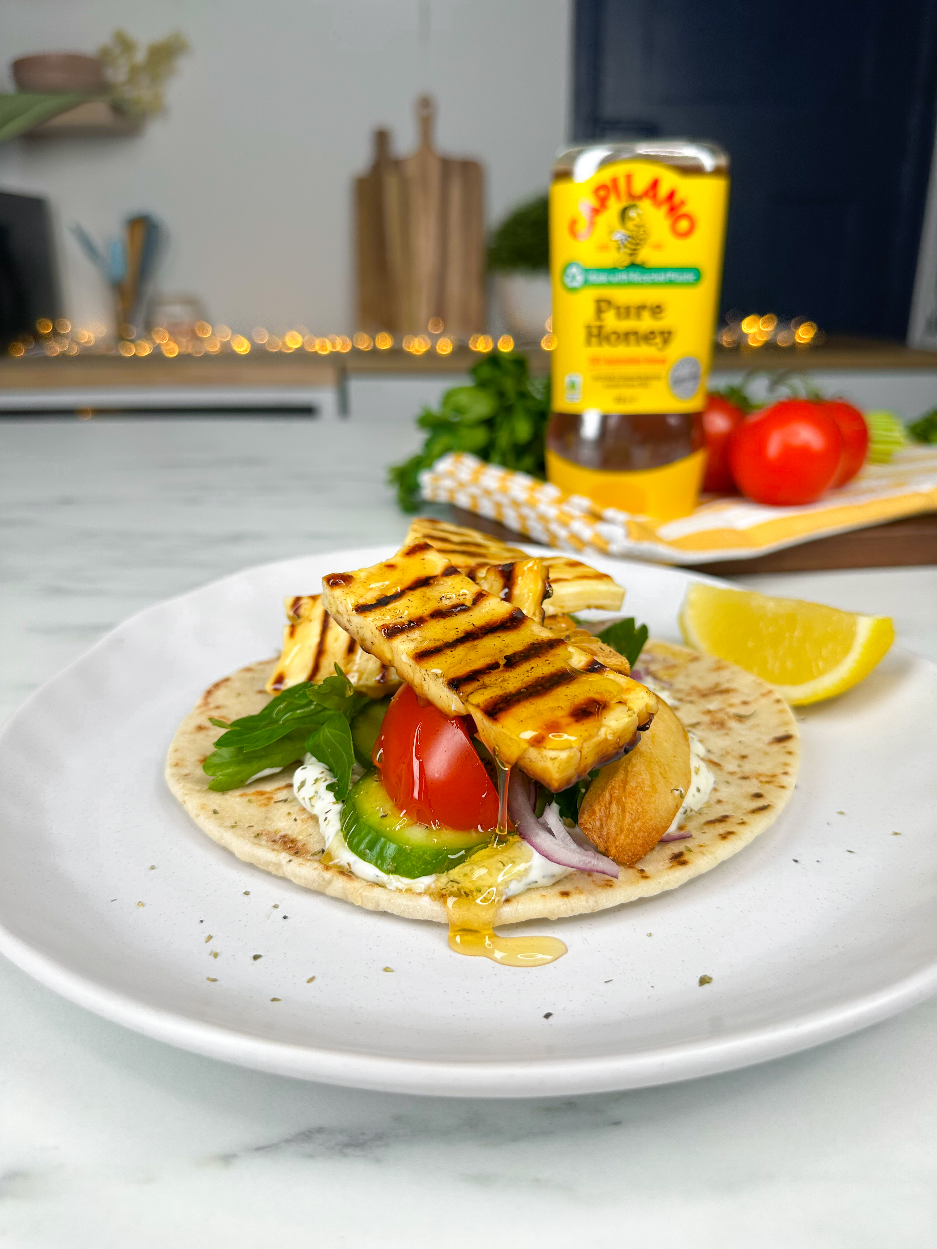 A grilled halloumi wrap is served on a white plate, with salad ingredients and honey drizzled on top. A bottle of Capilano Pure Honey is in the background