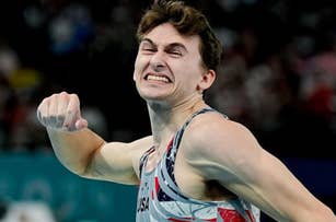 An Olympic gymnast clenches his fist and makes a determined face during a competition, wearing a gray and red uniform with USA on it