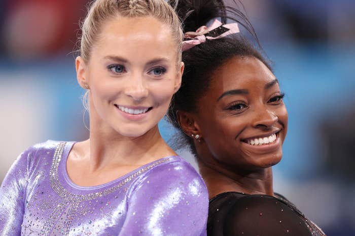 Two gymnasts, one in a sparkling outfit and the other in a stylish leotard, smile and pose back-to-back at a gymnastics event