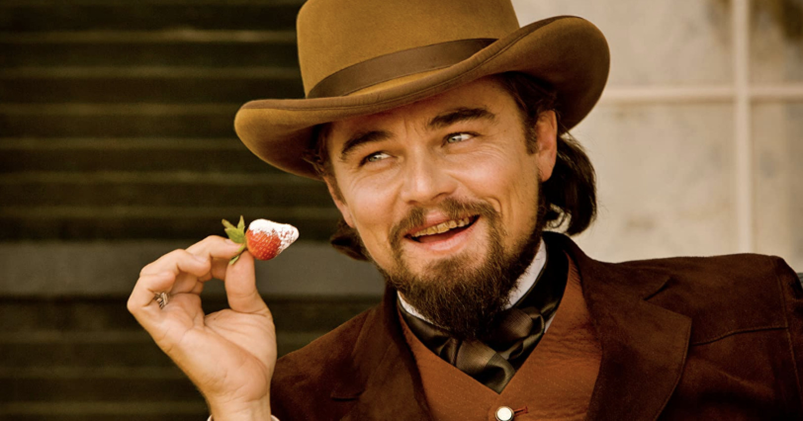 Leonardo DiCaprio in a period costume, holding a strawberry with a smile. He is wearing a brown hat and suit with a bow tie