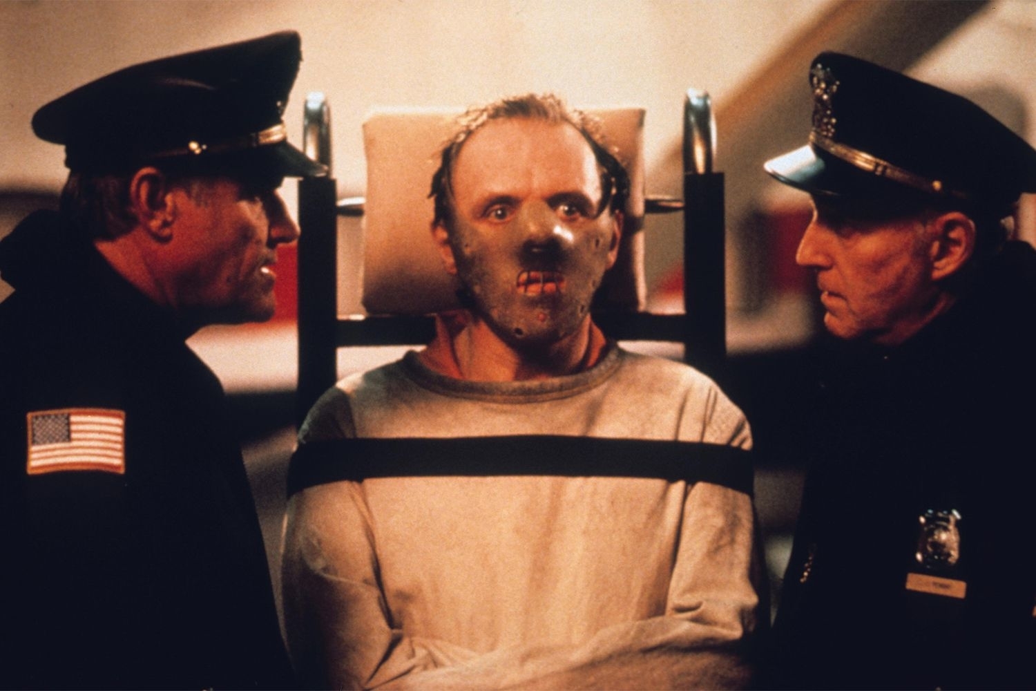 Two officers stand on either side of a restrained man wearing a mask in a scene from "The Silence of the Lambs," featuring Anthony Hopkins as Hannibal Lecter