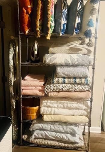 Metal shelving portion filled with assorted folded blankets, quilts, pillows, an iron, and a tiny yellowish basket, located successful a well-lit country with woody flooring