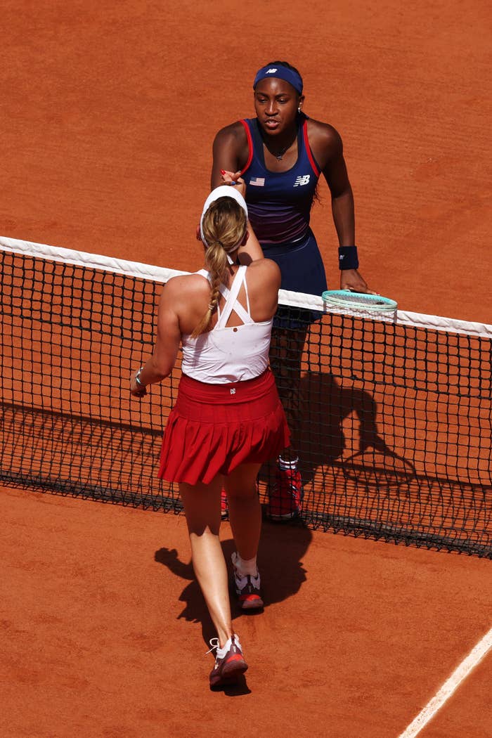 Coco Gauff and another tennis player shake hands at the net on a clay tennis court after a match