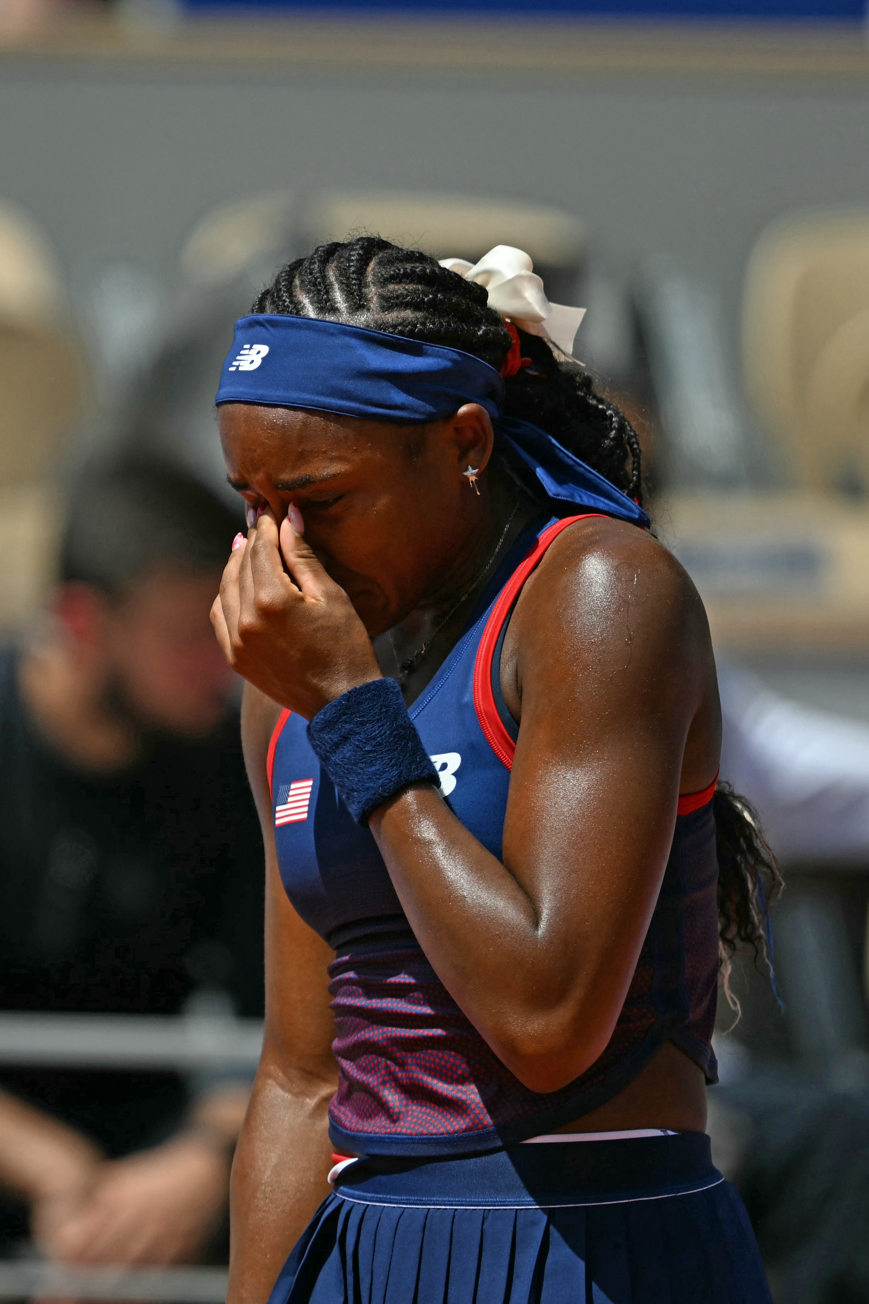 Coco Gauff is on a tennis court, wiping away tears while wearing a sports headband and a sleeveless sport top with her hair in braids