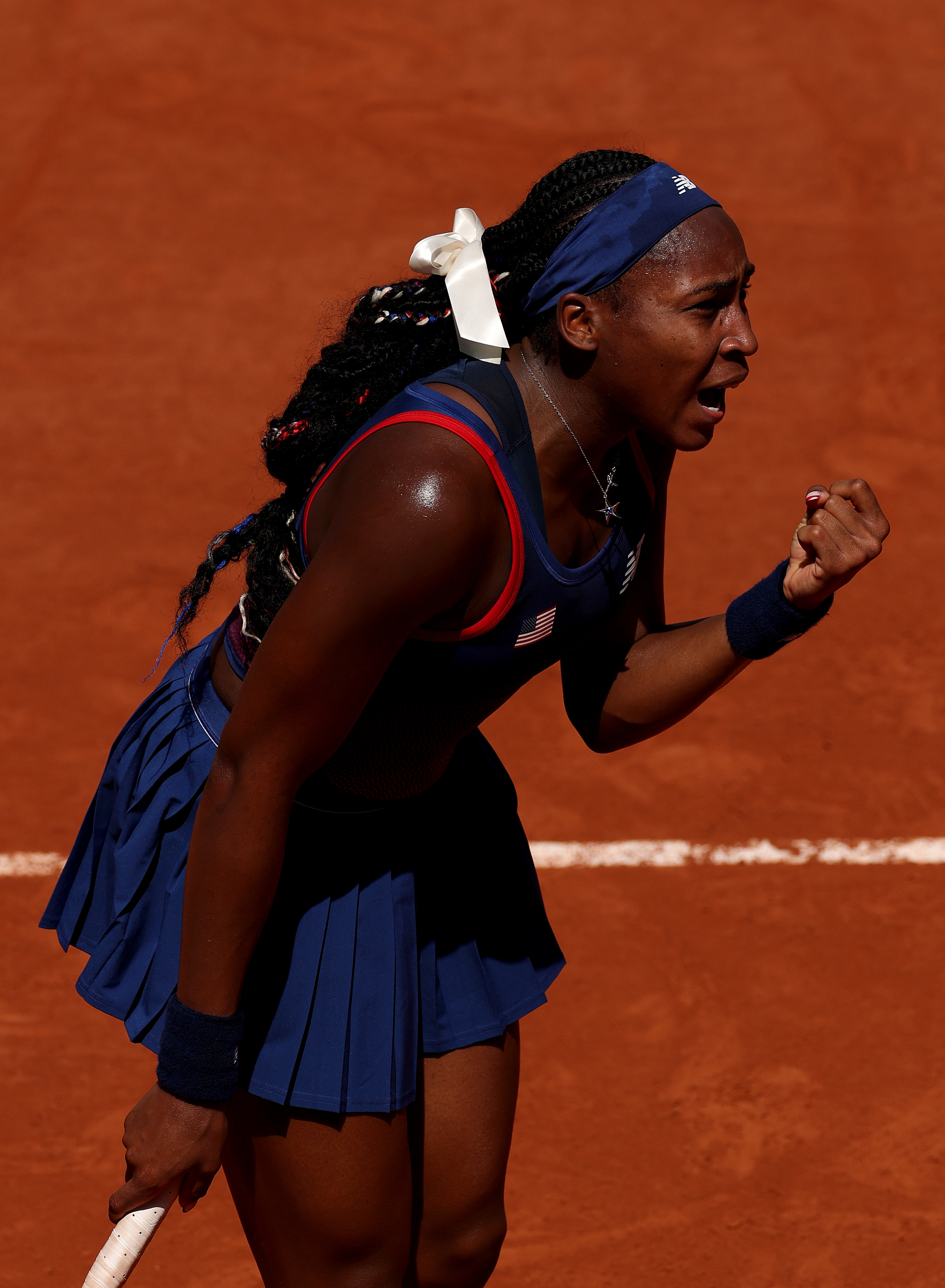 Coco Gauff celebrates a tennis victory on a clay court, holding her racket, wearing a navy sports dress and headband with a white bow