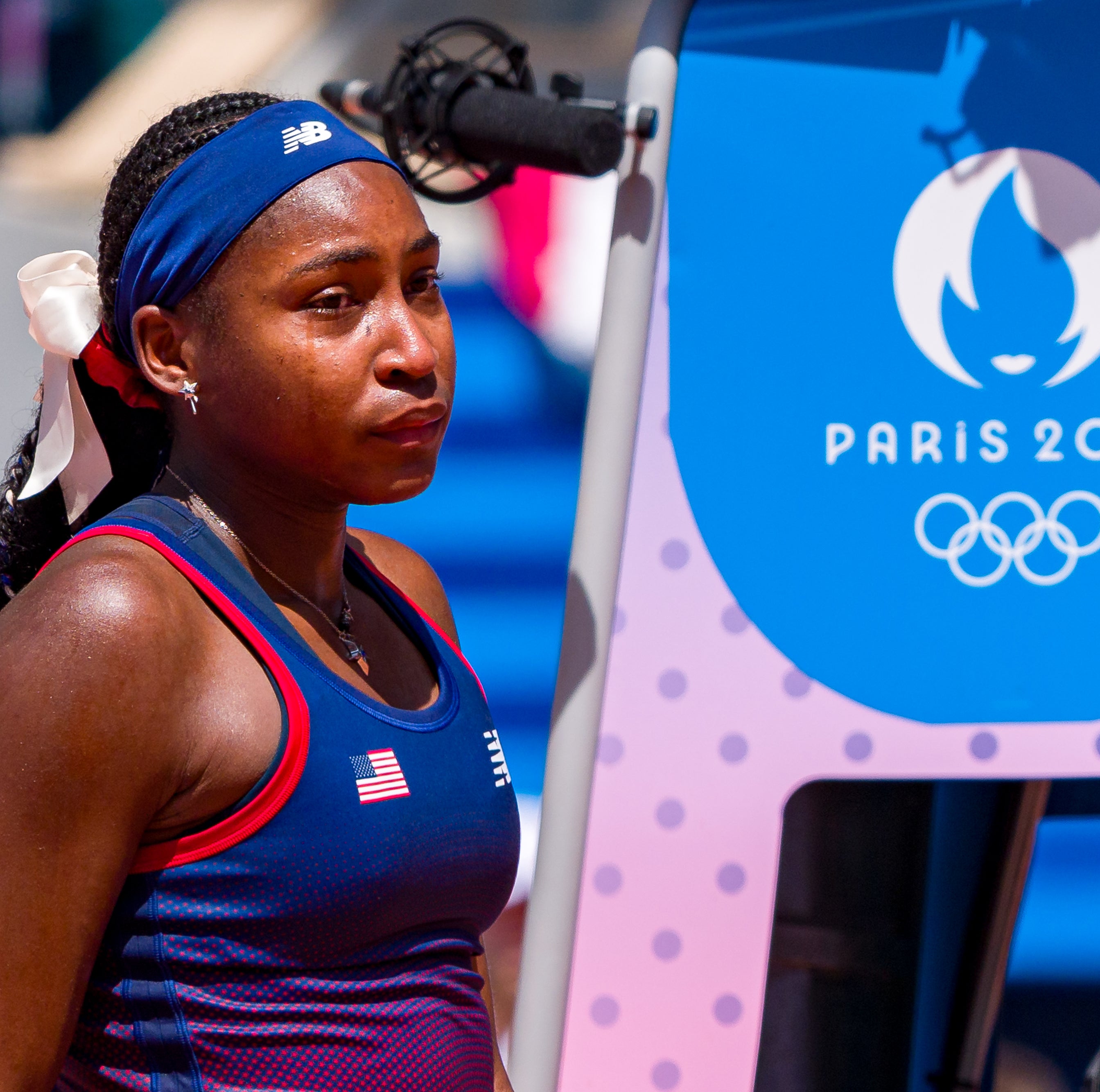 Coco Gauff stands on a tennis court, wearing a sports outfit with a headband and a ribbon in her hair, next to a sign for Paris 2024 Olympics