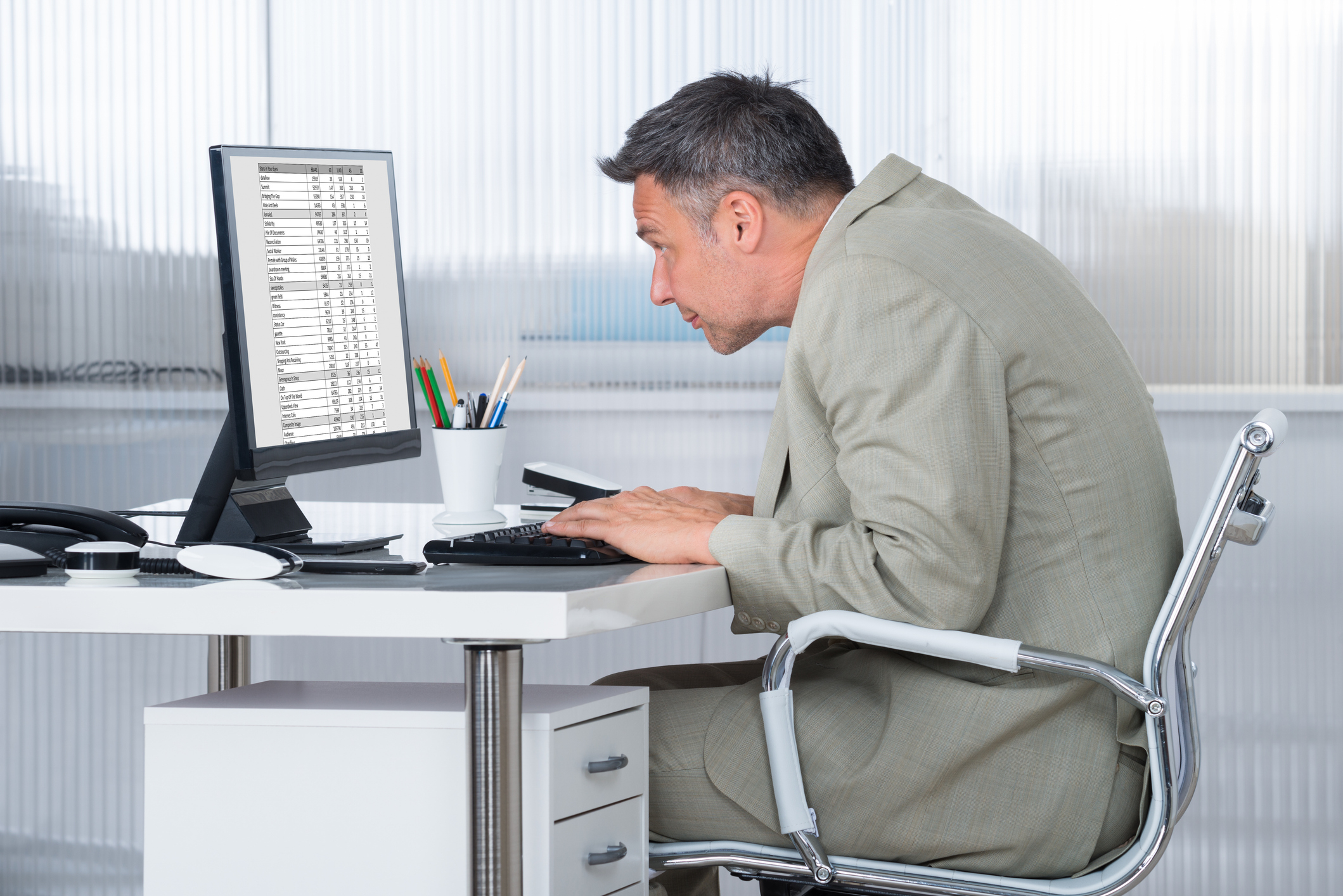 A man in a suit is sitting at an office desk, leaning in close to a computer monitor with a spreadsheet on the screen.
