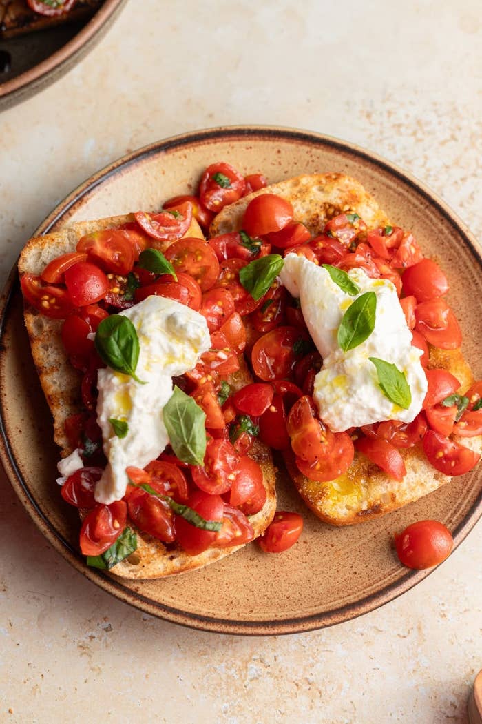 Two slices of bread topped with chopped tomatoes, basil leaves, and dollops of creamy cheese on a plate