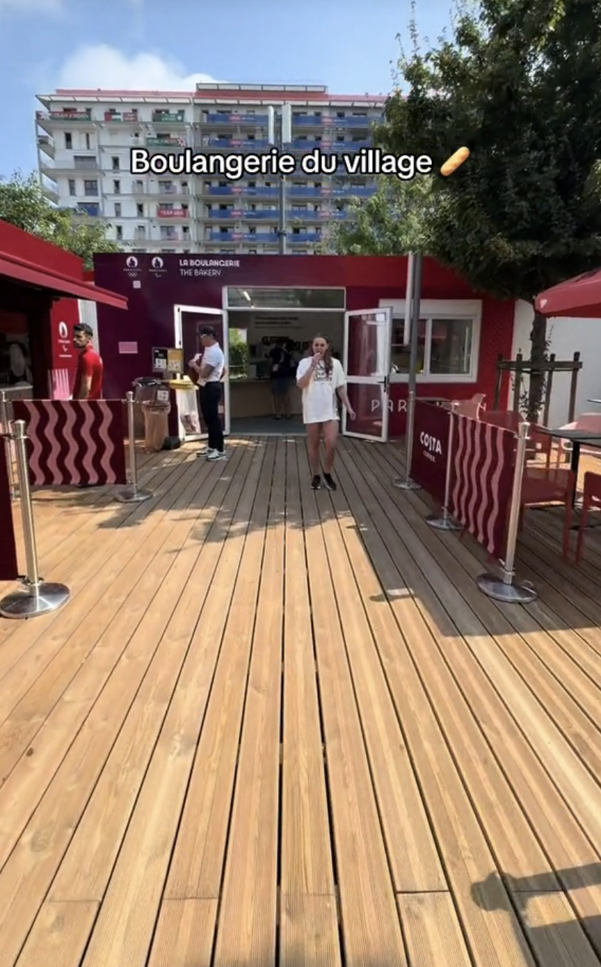 People standing outside Boulangerie du village, a bakery, with outdoor seating and a wooden deck
