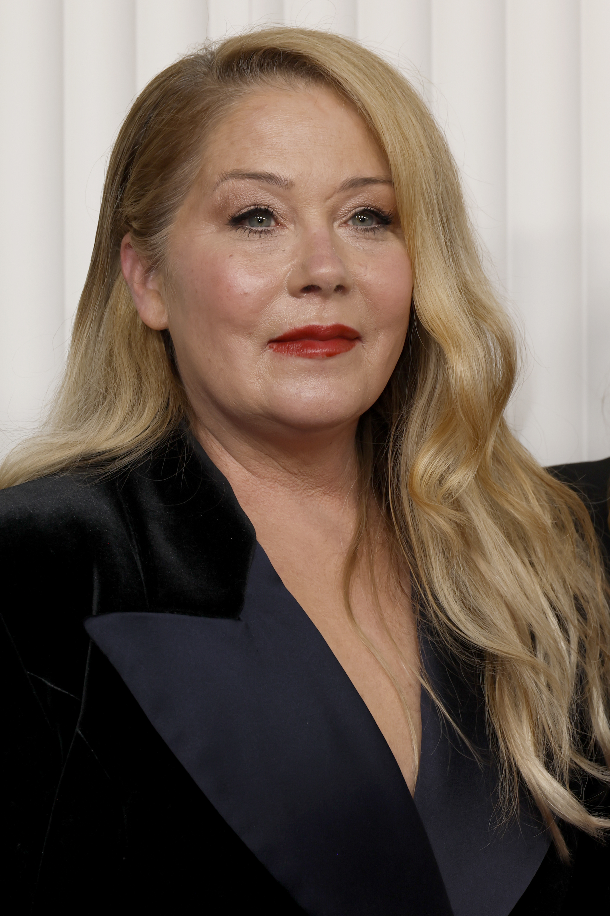 Christina Applegate is pictured in a sophisticated black suit jacket with long, wavy hair cascading over her shoulder, standing in front of a neutral background