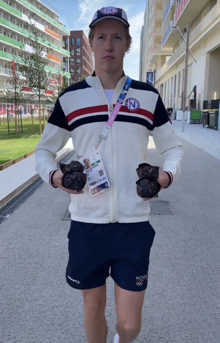 Person in casual athletic clothing, wearing a cap and ID badge, holds two bouquets of flowers. Buildings and greenery are in the background