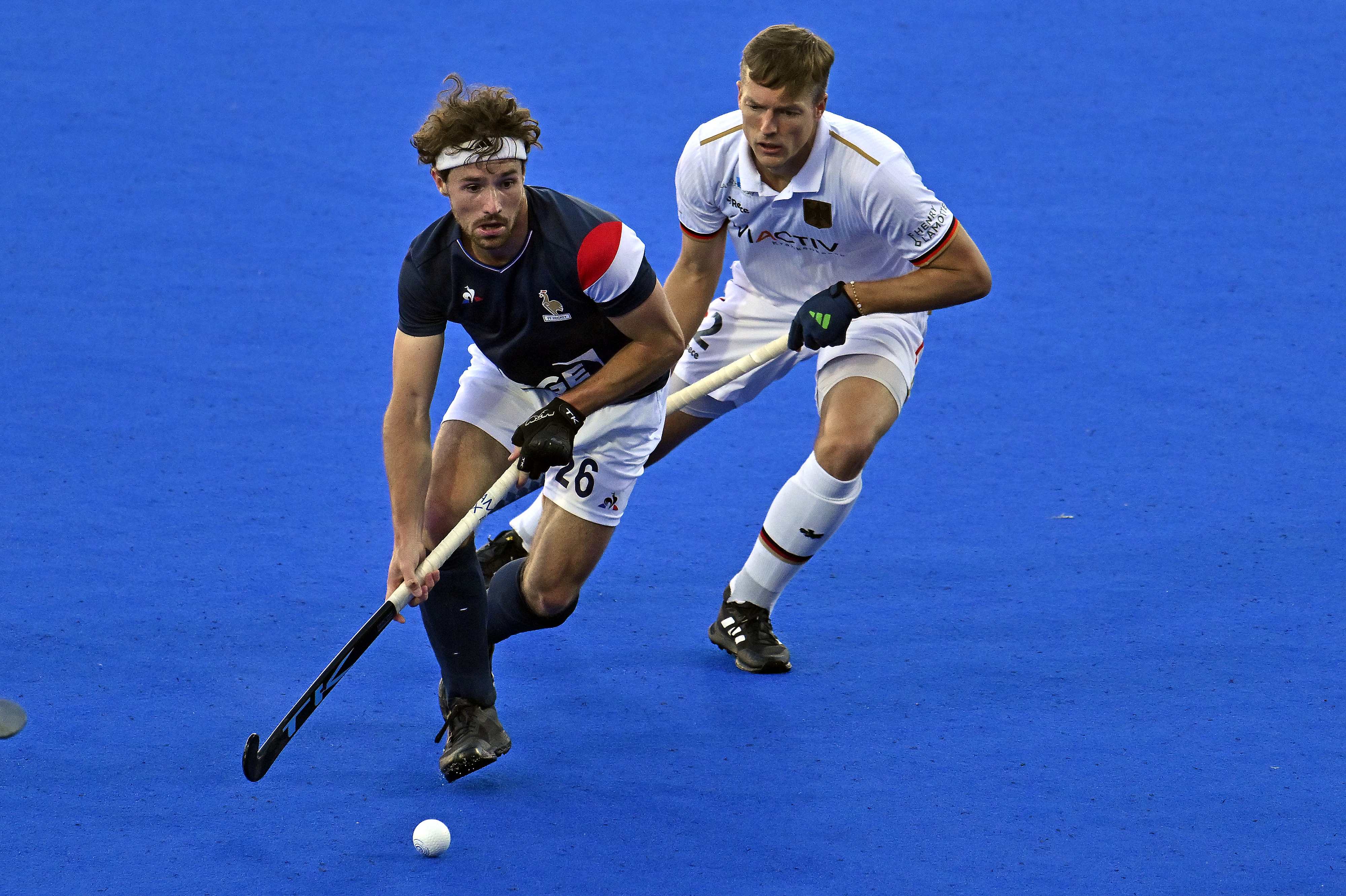 Two of France and Germany's field hockey players in action on the blue pitch during a match, one about to hit the ball while another defends closely behind