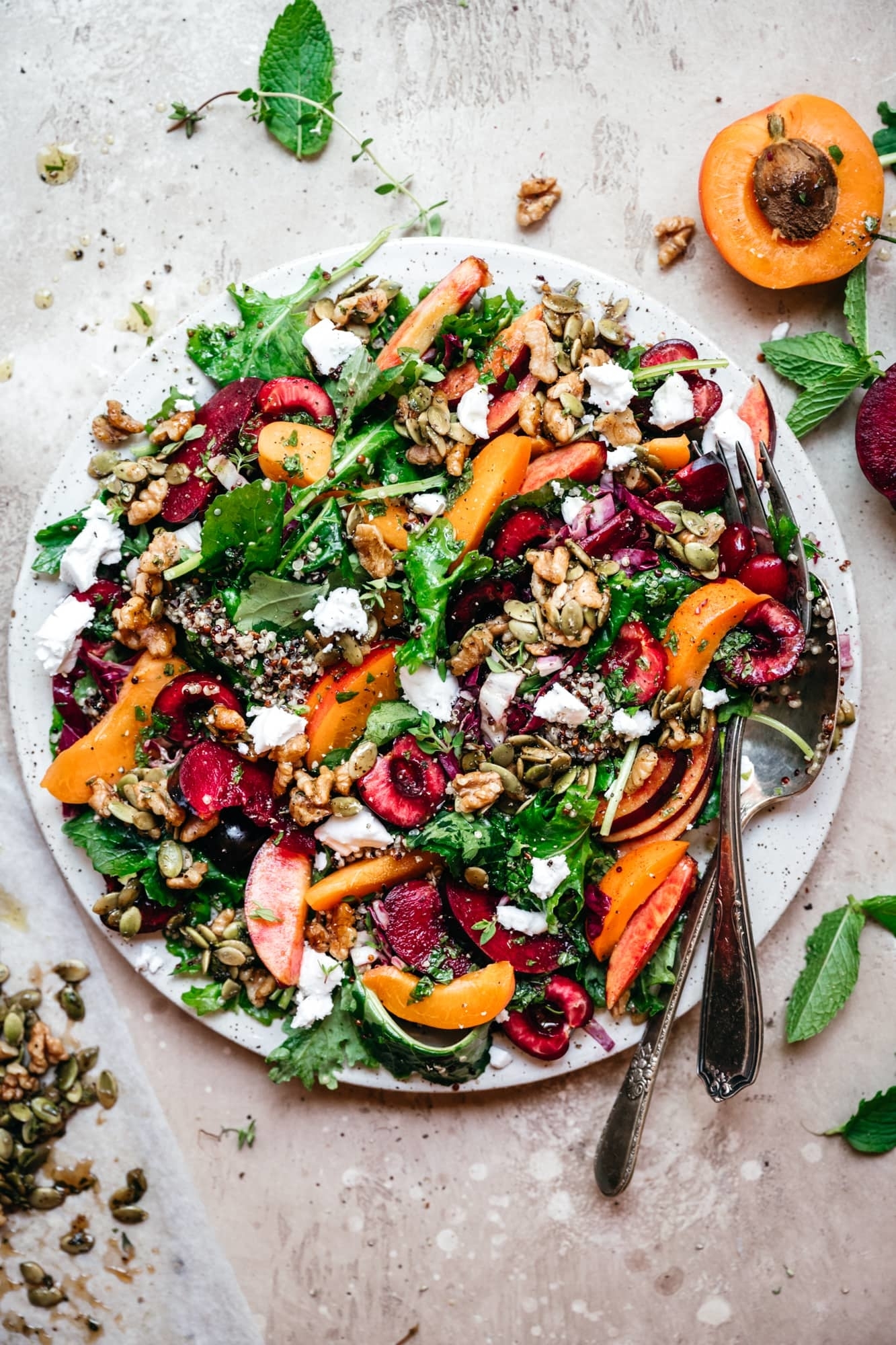 A fresh summer salad with mixed greens, sliced peaches, cherries, walnuts, and crumbled cheese, accompanied by a fork and spoon on the side