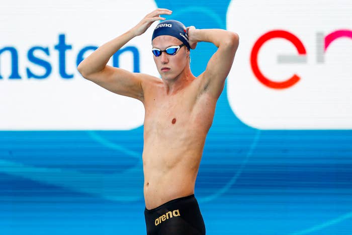 A male swimmer in black swimwear and a swim cap adjusts his goggles before a race