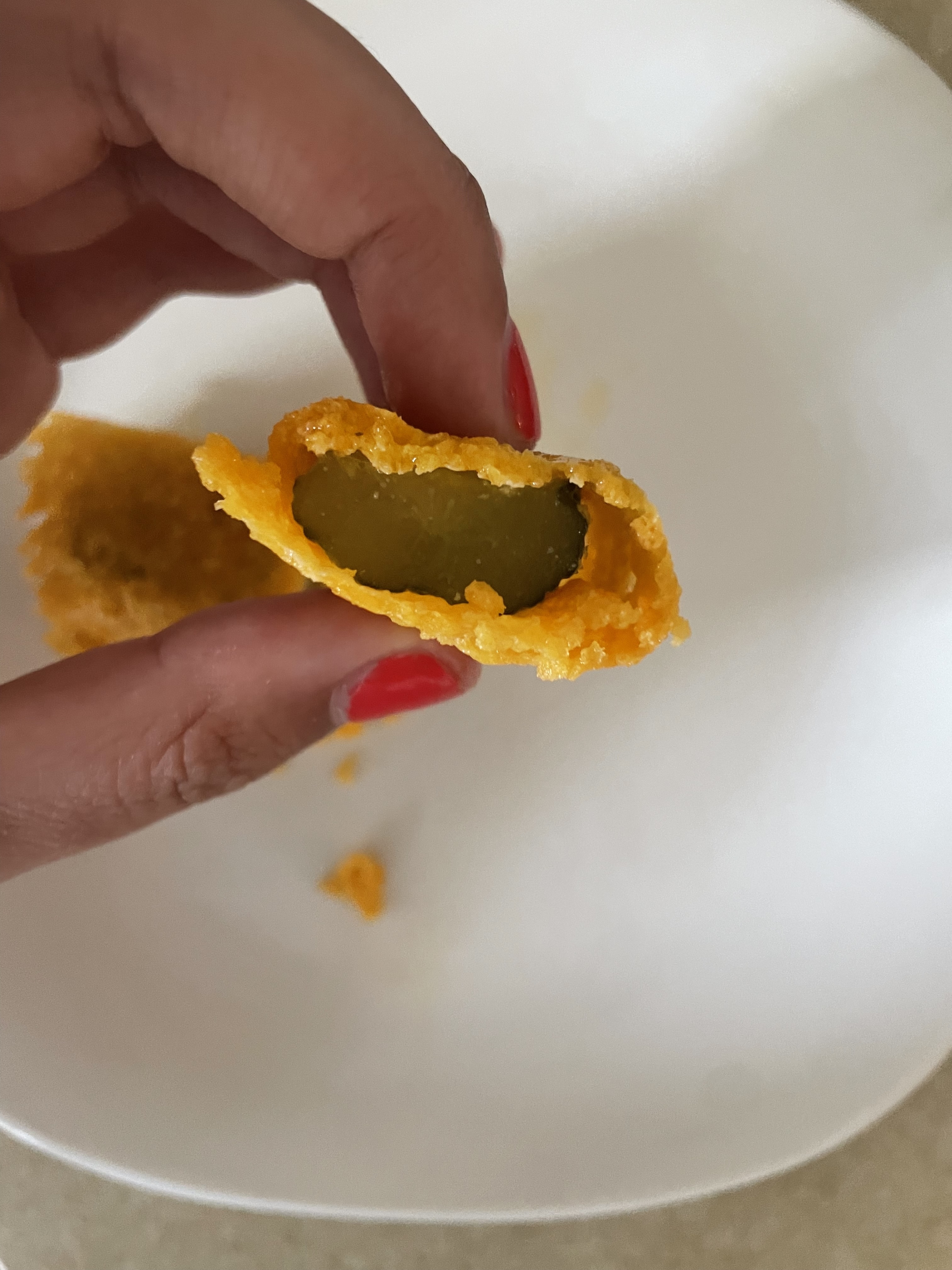 A hand with red-painted nails holds a piece of fried food with a pickle inside on a white plate