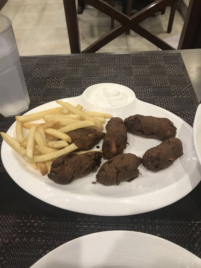 A plate of French fries, fried sausage rolls, and a dollop of white sauce on a woven placemat at a restaurant table. A glass of water is beside the plate