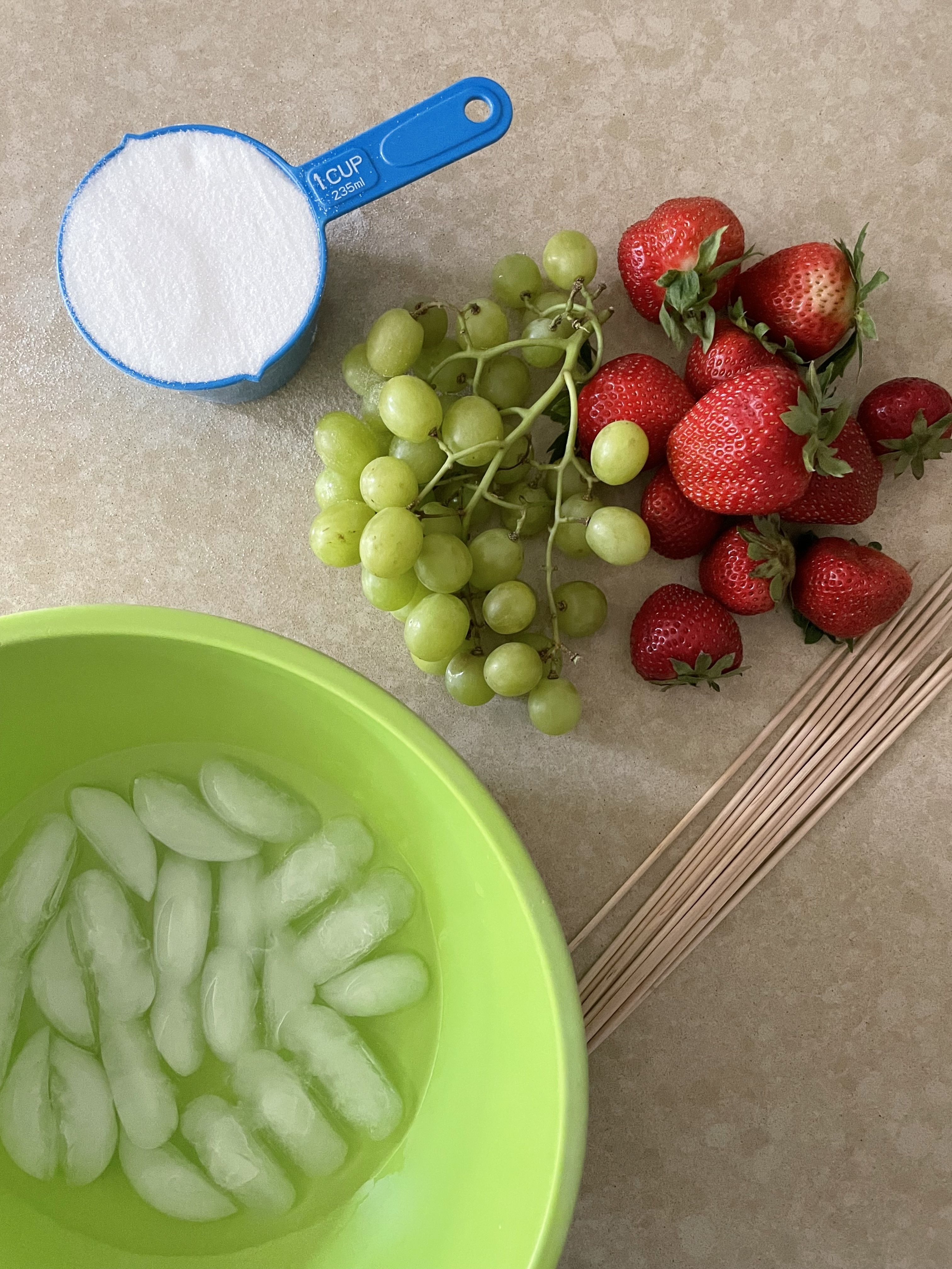 A bowl of ice water, a cup of sugar, green grapes, and strawberries are arranged on a countertop with wooden skewers beside them