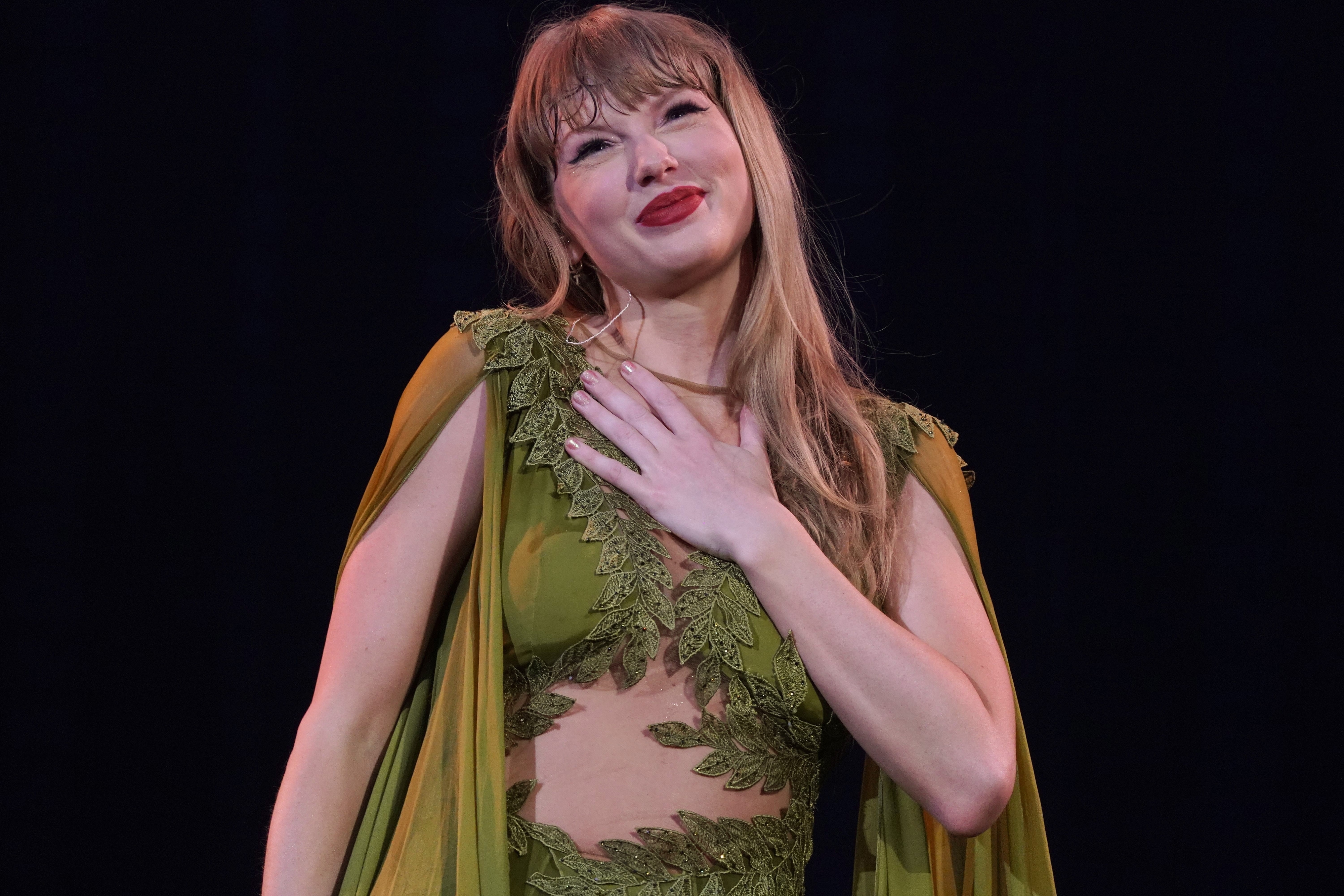 Taylor Swift smiles with one hand on her chest, wearing a stylish dress with intricate leaf designs