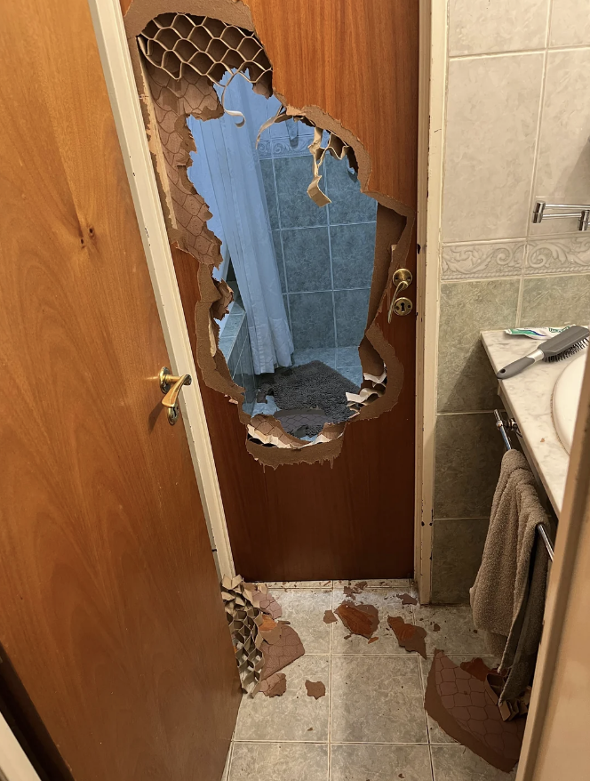 A wooden door with a large, jagged hole smashed through it, revealing a bathroom with tiled walls and a towel hanging on a rack