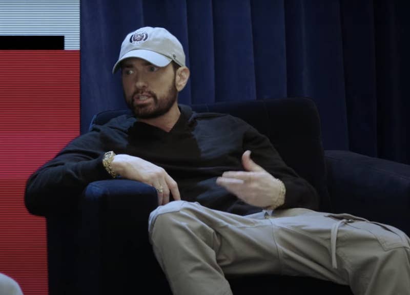 Eminem is seated on a couch, wearing a baseball cap, black long-sleeve shirt, and beige pants, discussing a topic with a visible hand gesture