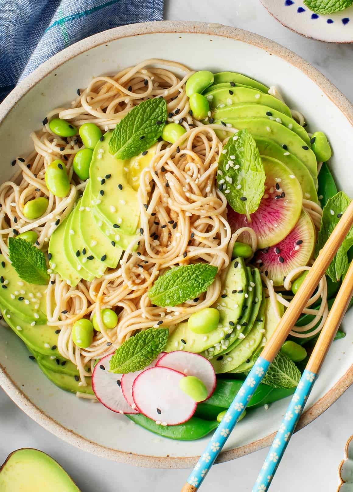 A bowl of soba noodles topped with avocado slices, edamame, radish slices, and mint leaves, garnished with sesame seeds, with a pair of chopsticks on the side