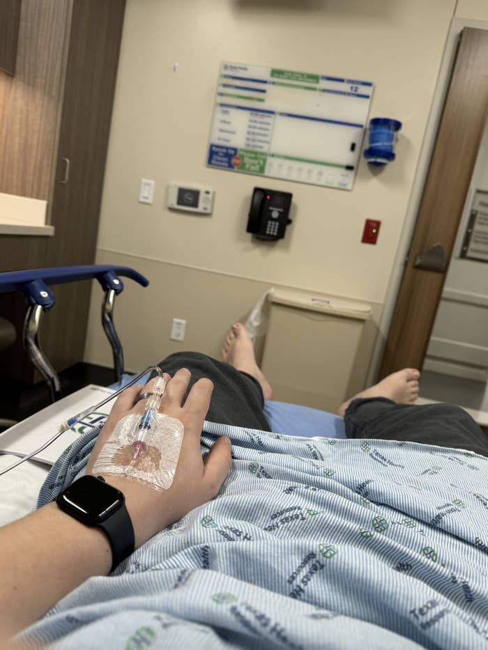 A person lies on a hospital bed wearing a wristwatch. Their hand has an IV line attached. The room has medical equipment and a whiteboard in the background