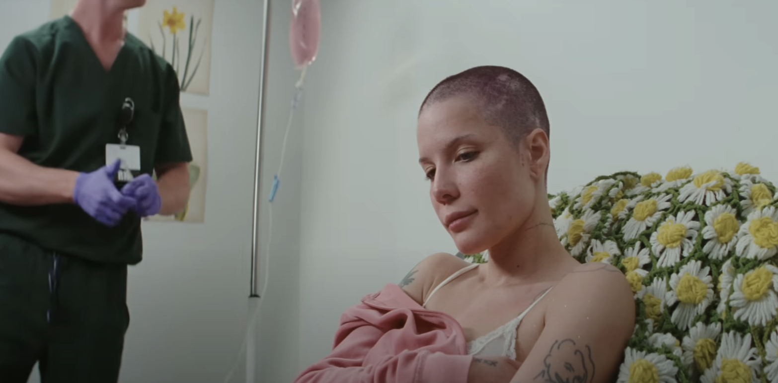 Halsey with shaved head sits on a chair with a floral blanket in a hospital room. A medical professional in scrubs stands nearby