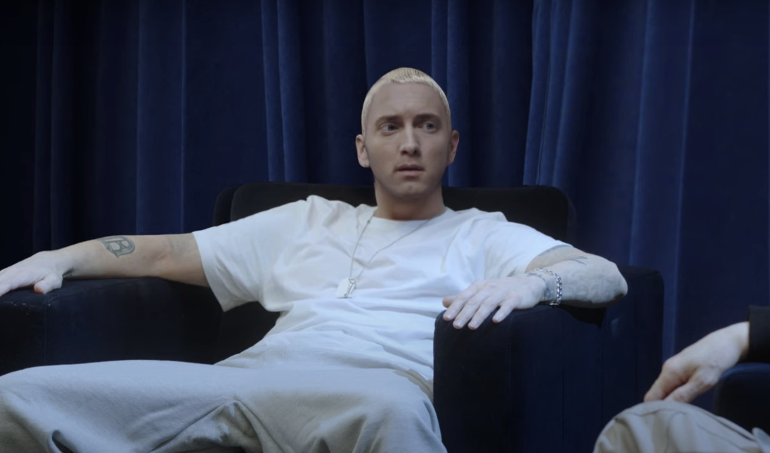 Eminem sits on a black chair with a blue curtain background, wearing a white t-shirt and light pants, with visible tattoos and a chain necklace