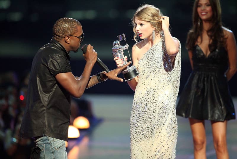 Kanye West interrupts Taylor Swift, holding a trophy in a sparkly dress, during the MTV Video Music Awards. Another woman in a black dress stands in the background