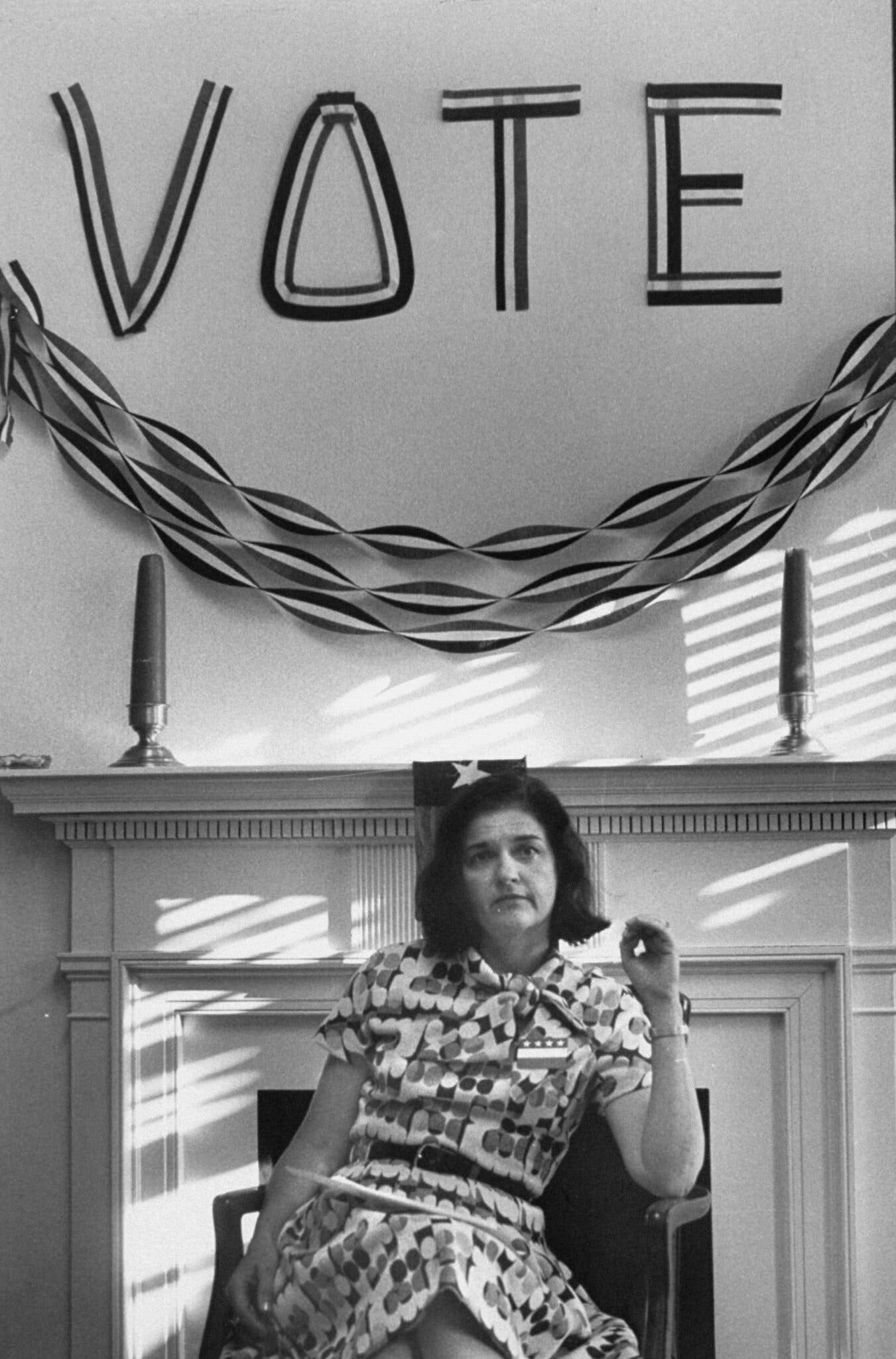 Sissy Farenthold in a patterned dress sits under a "VOTE" sign with ribbons on a mantel