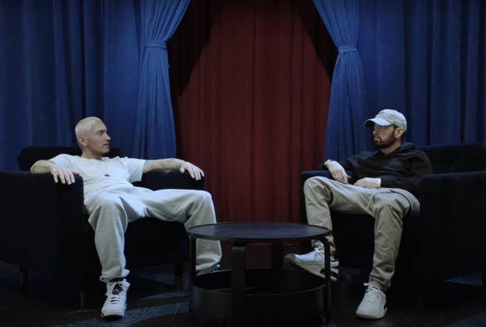 Eminem and Eminem sit on chairs facing each other in a relaxed setting. One wears a white shirt and pants, the other a black hoodie, khaki pants, and a beige cap