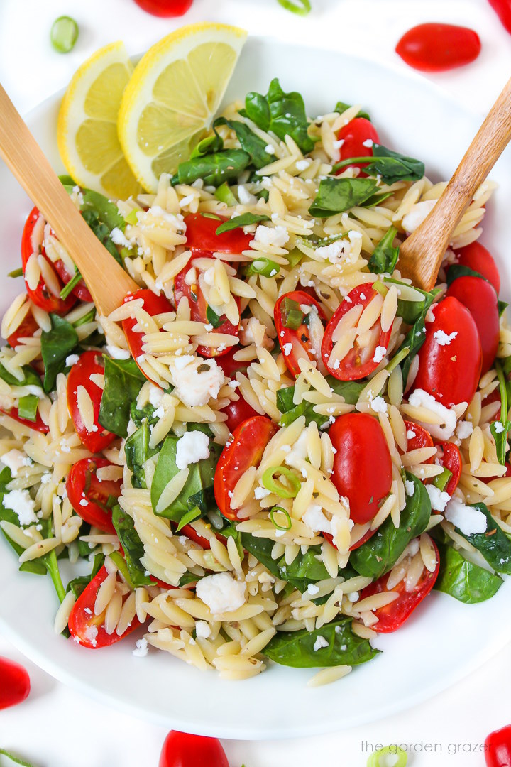 A bowl of orzo pasta salad with cherry tomatoes, spinach, feta cheese, and two lemon slices on the side. Two wooden spoons are mixing the salad