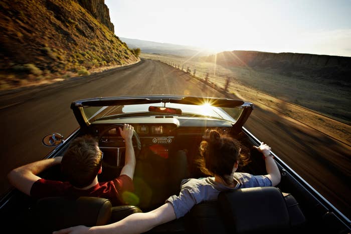 Two people are driving on a scenic, winding road at sunset in a convertible; the person on the right has their arm across the back of the front seat