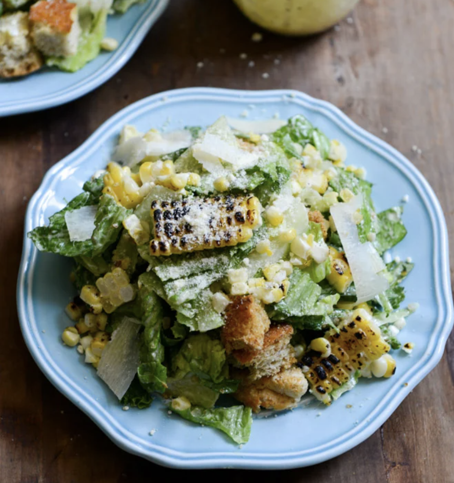 A fresh Caesar salad on a blue plate, topped with croutons, grilled corn, shaved Parmesan, and creamy dressing