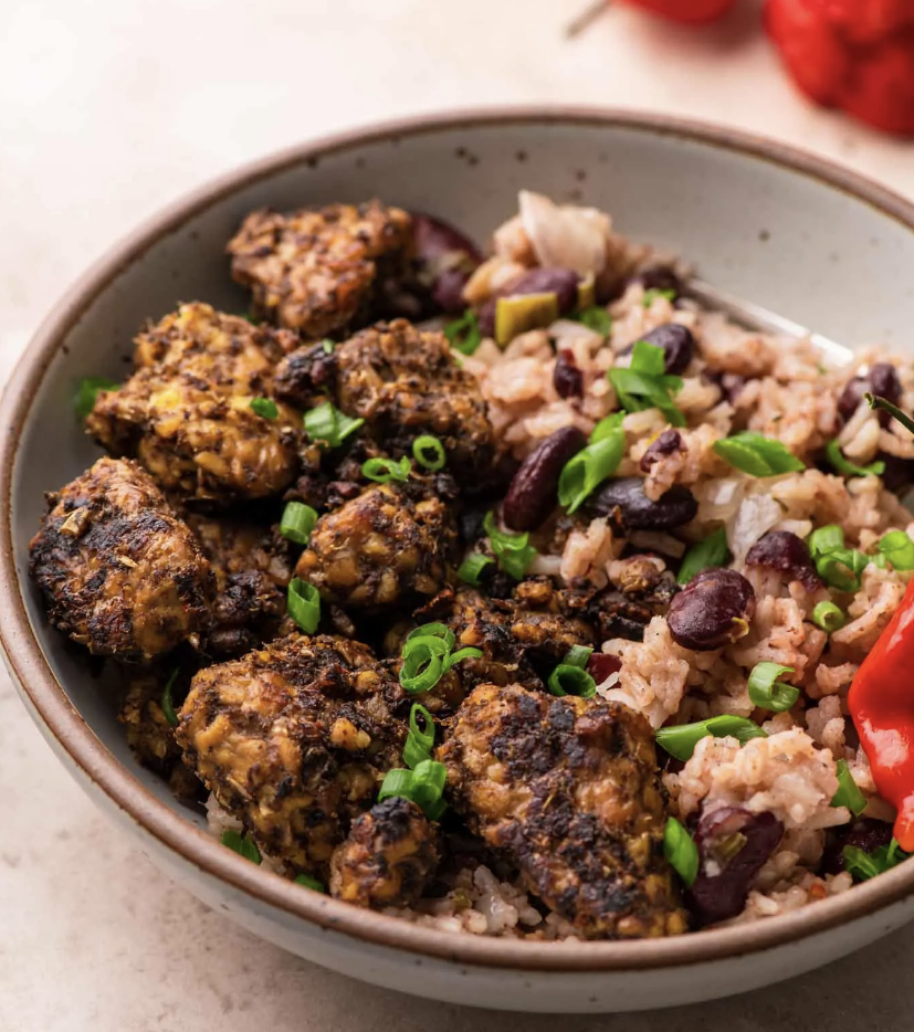 A bowl of rice and kidney beans topped with chunks of seasoned, grilled meat and garnished with chopped green onions and a red pepper