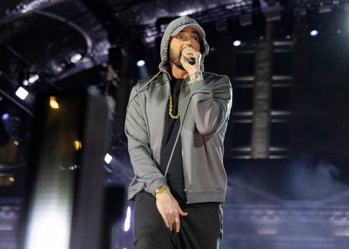 Eminem performs on stage wearing a gray hoodie, gold chain, and black pants, holding a microphone