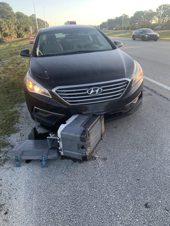 A black Hyundai sedan is on the roadside, its front partially damaged by a black and gray box-like object lodged under the bumper. Another car is on the road in the background