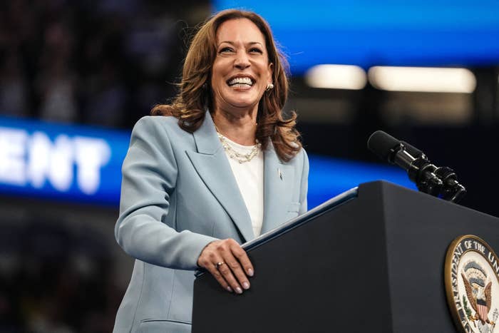 Kamala Harris stands smiling at a podium with microphones, wearing a blazer and a white top. She is giving a speech at an event