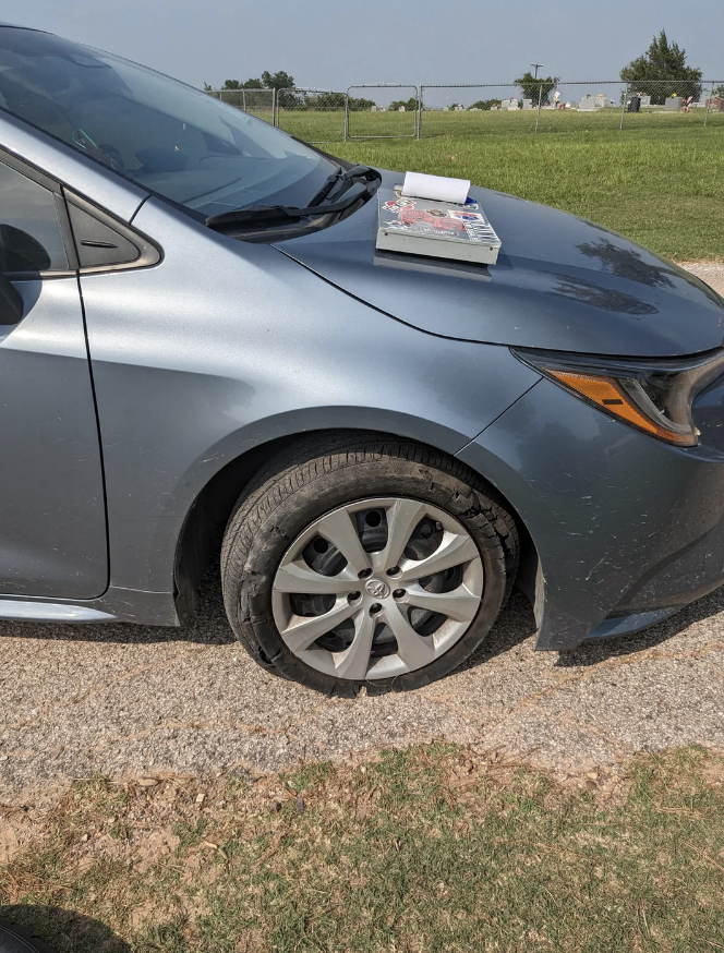 A car with a flat front tire on the side of a road. Two books rest on the car's hood