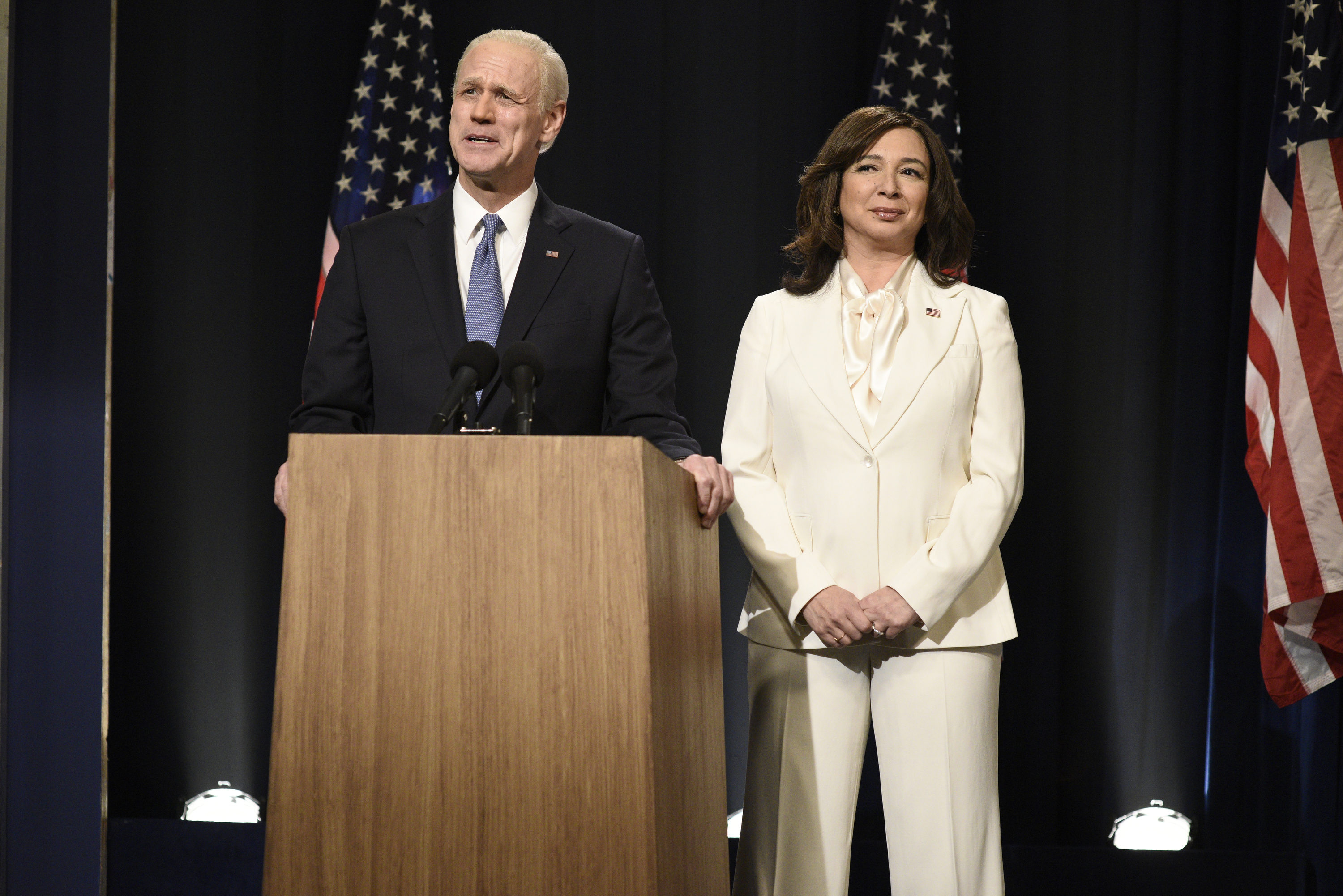 Two people stand at a podium in front of American flags. The man is in a suit, and the woman, Maya Rudolph, wears a white suit