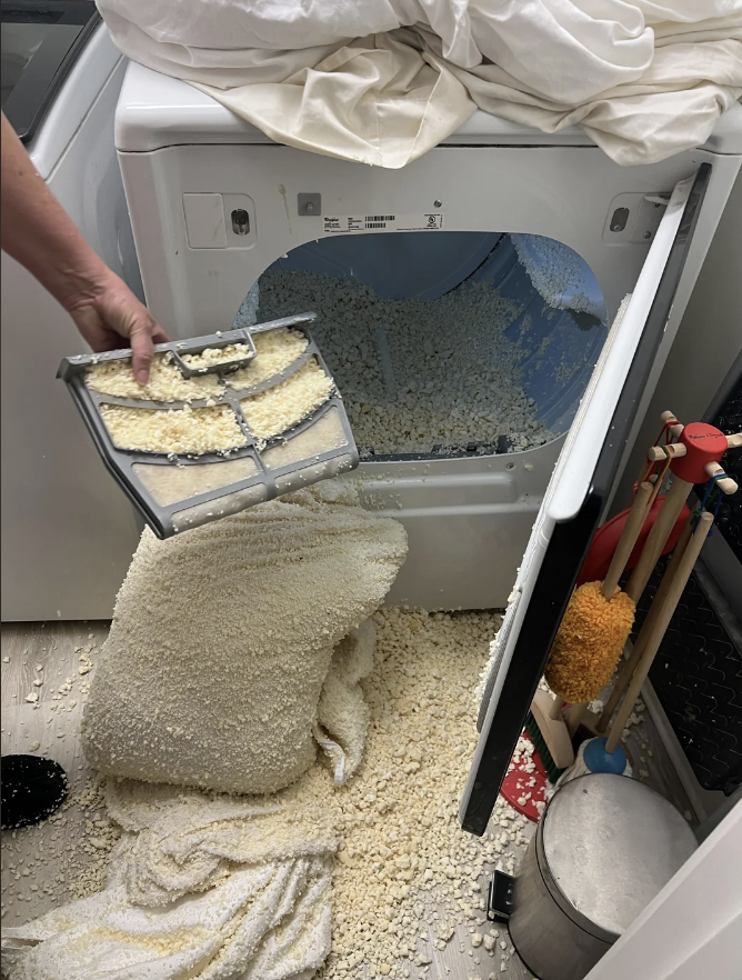 A person holding a clogged lint filter in front of a dryer spilling wet shredded foam pieces onto the floor, a pillow, and a shelf with cleaning supplies nearby