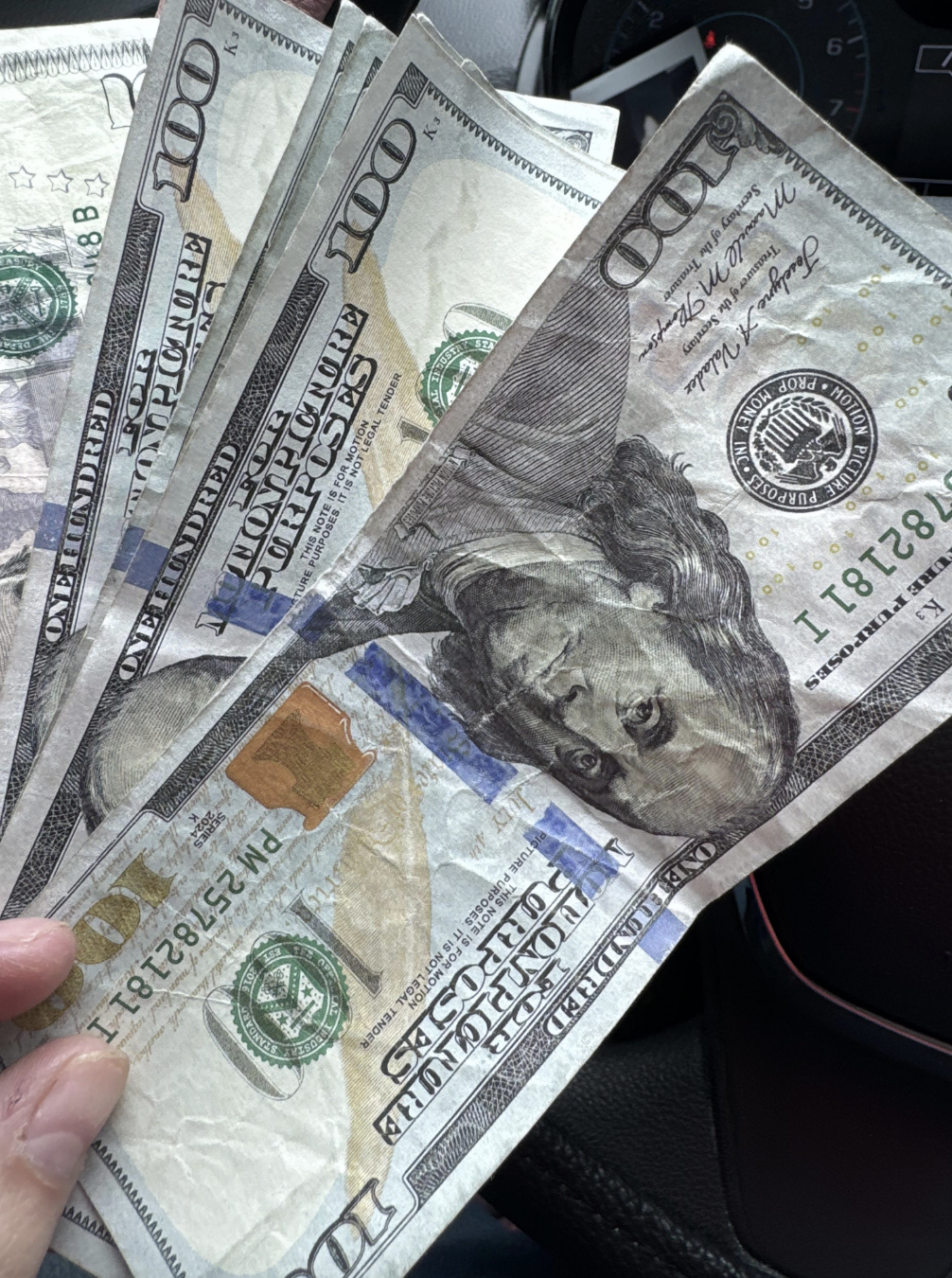 A hand holds several $100 bills. The background includes part of a car dashboard
